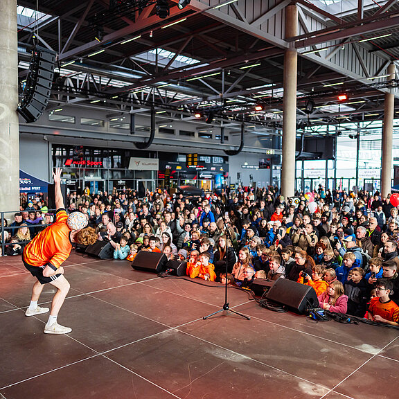 Ein Fußballer in orangenem Trikot steht auf einer Bühne und zeigt den Kindern Tricks mit einem Fußball
