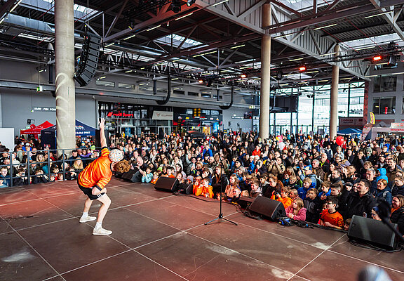 Ein Fußballer in orangenem Trikot steht auf einer Bühne und zeigt den Kindern Tricks mit einem Fußball