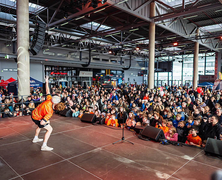 Ein Fußballer in orangenem Trikot steht auf einer Bühne und zeigt den Kindern Tricks mit einem Fußball